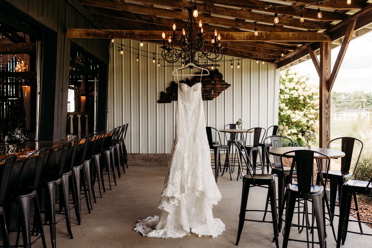 Elegant wedding dress hanging from a chandelier in a rustic barn venue with string lights and black metal chairs.