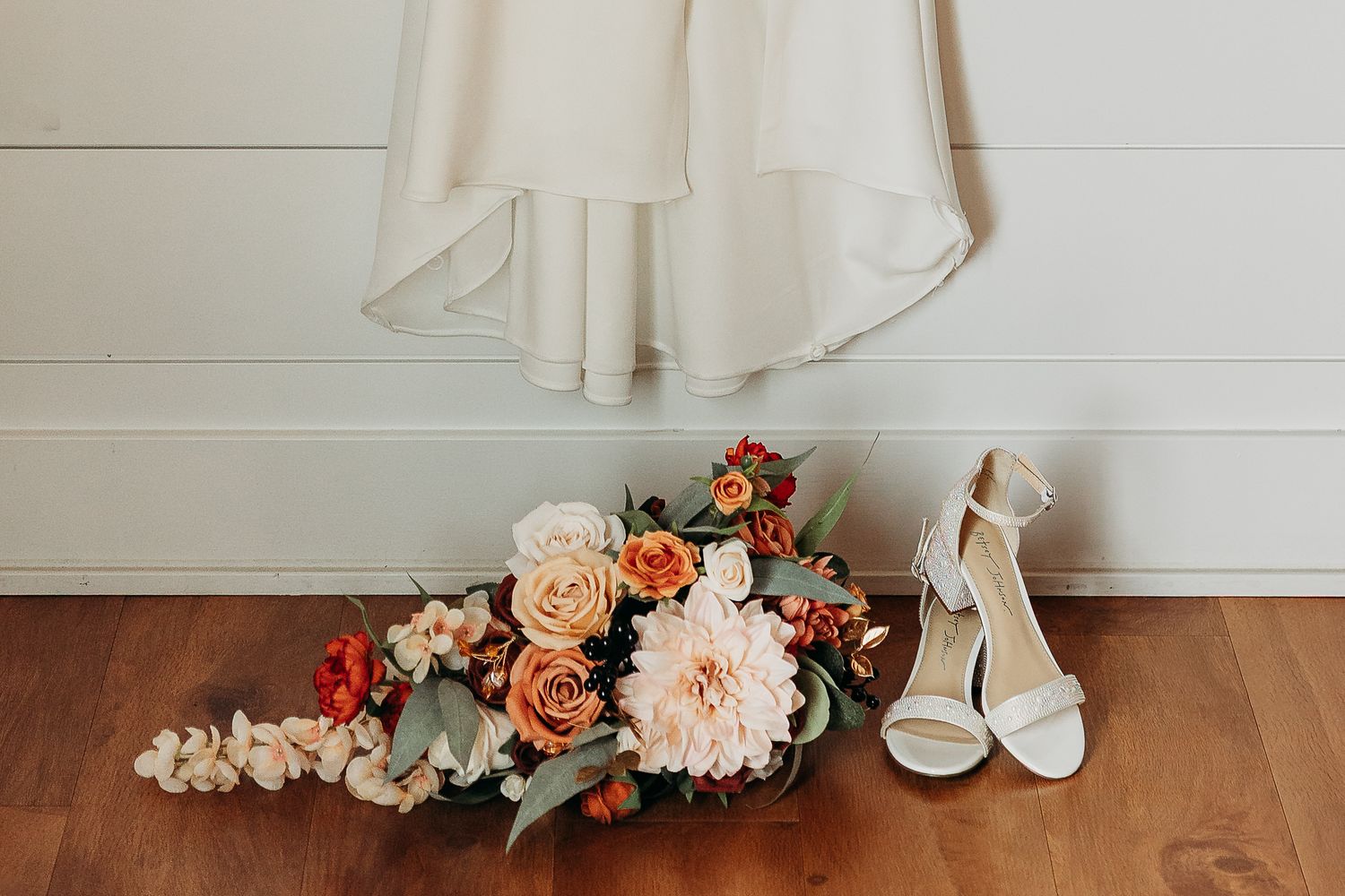 Wedding details flat lay featuring a white bridal gown, ivory heels, and a vibrant orange and pink floral bouquet on wood floor.