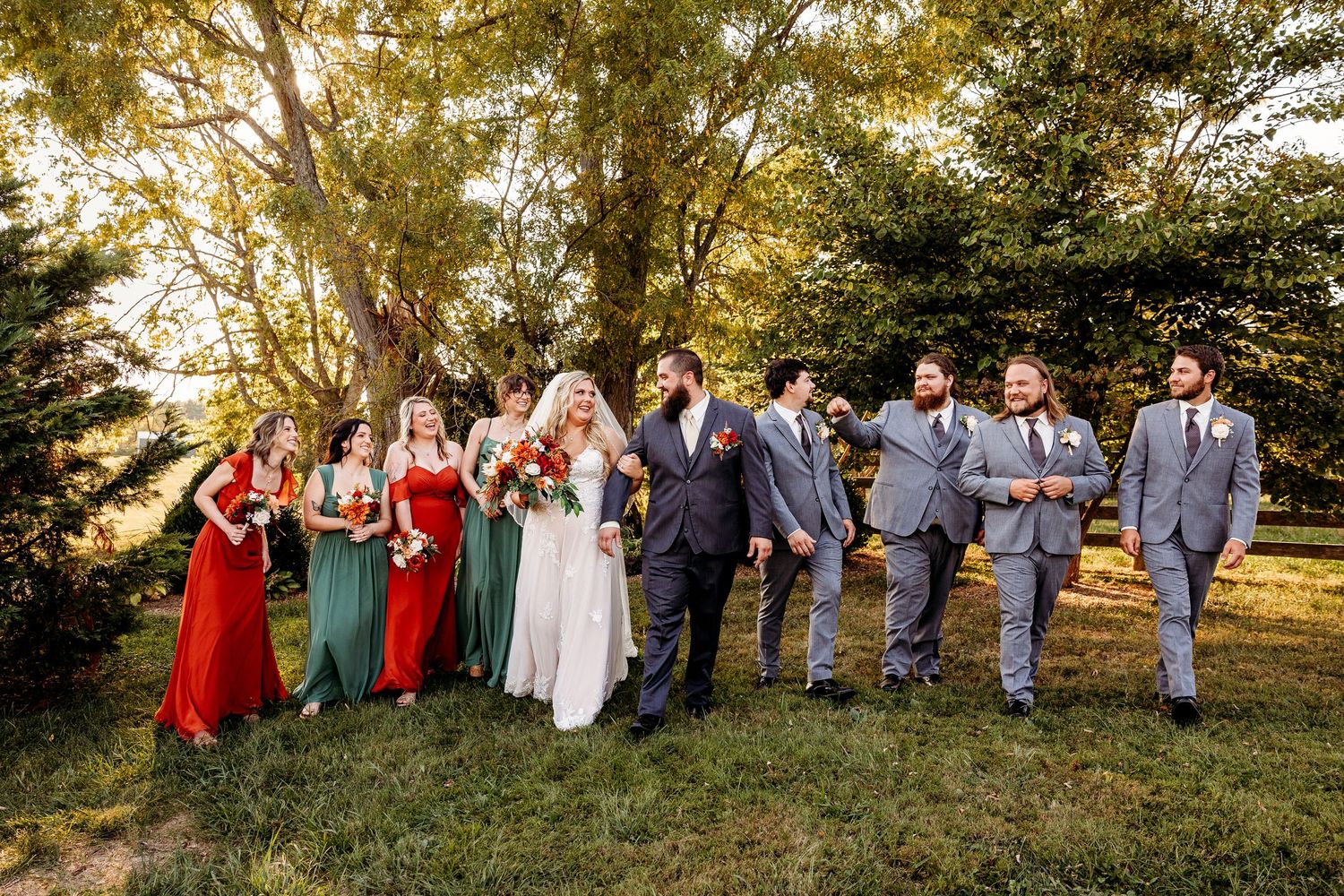 Wedding party in red dresses and gray suits poses on a grassy hill during golden hour.
