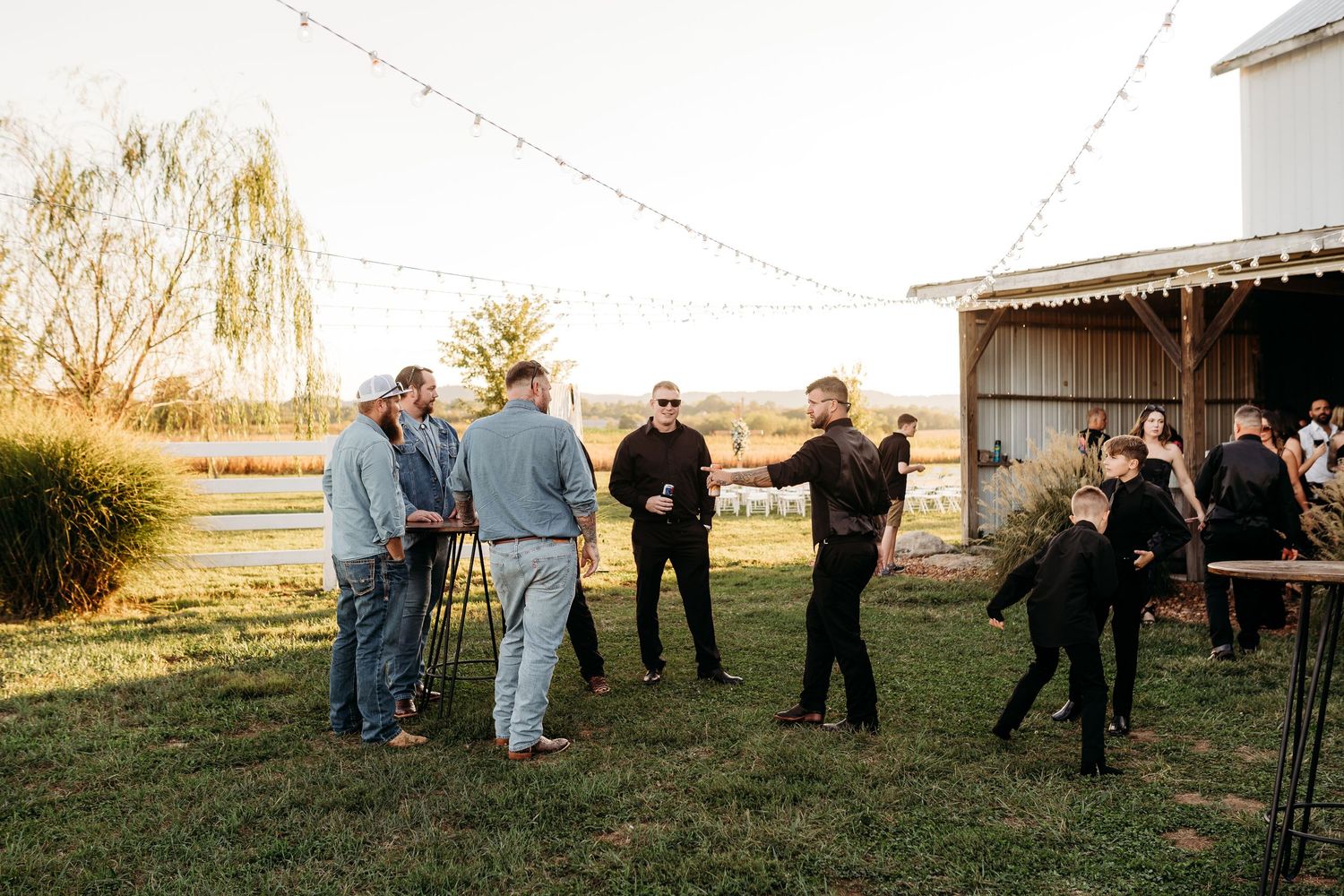 Wedding guests mingle outdoors near a white barn at golden hour with string lights overhead at sunset.