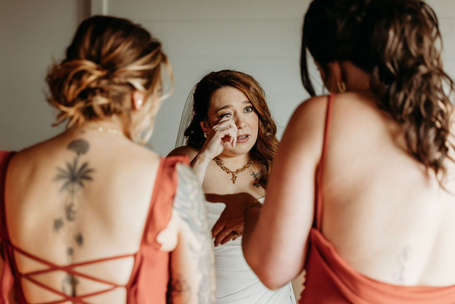 Emotional bride wipes tears while bridesmaids in rust-red dresses comfort her during wedding day preparations.