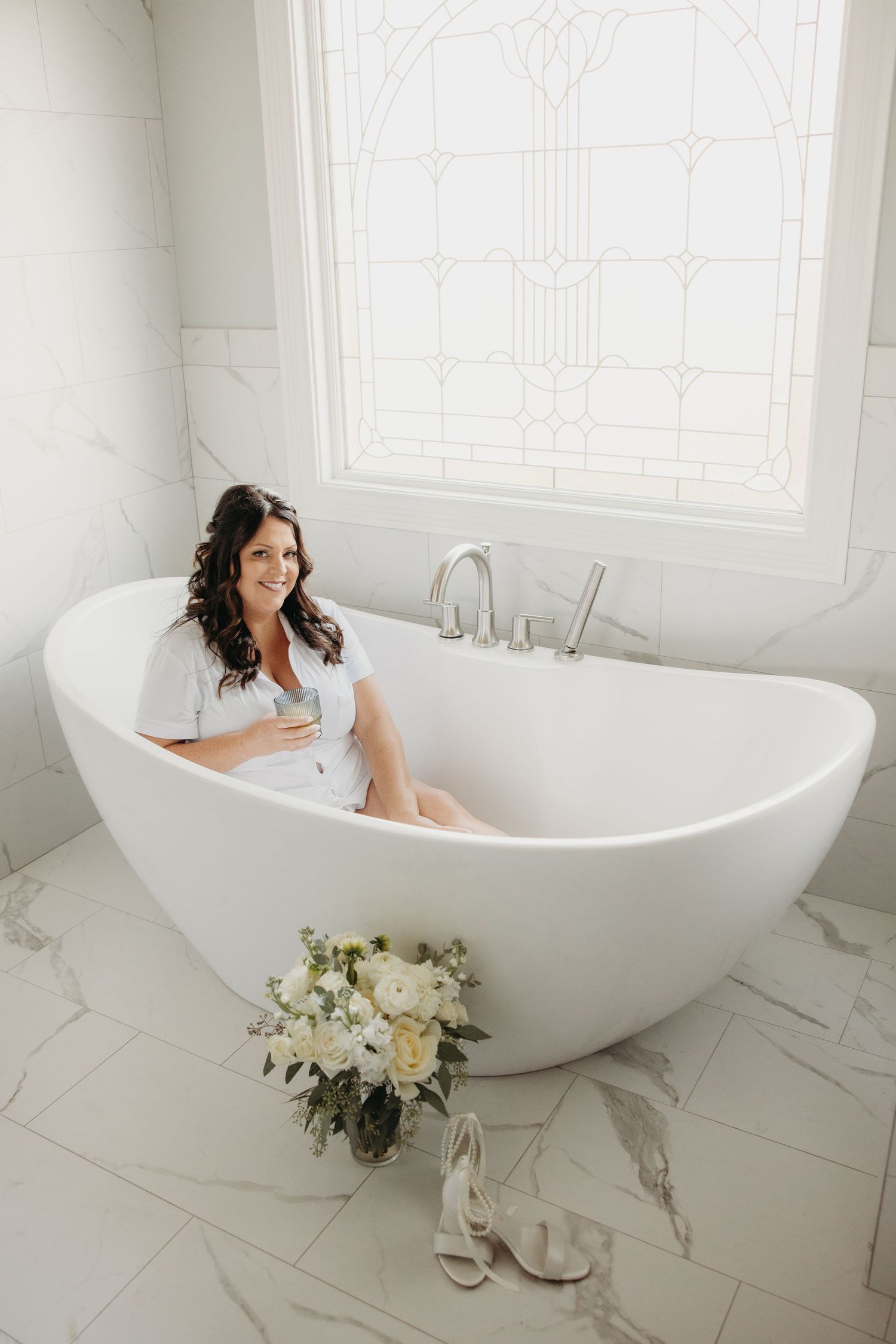 Bride sitting in a freestanding white bathtub holding a glass, with a bridal bouquet and heels on the marble floor.