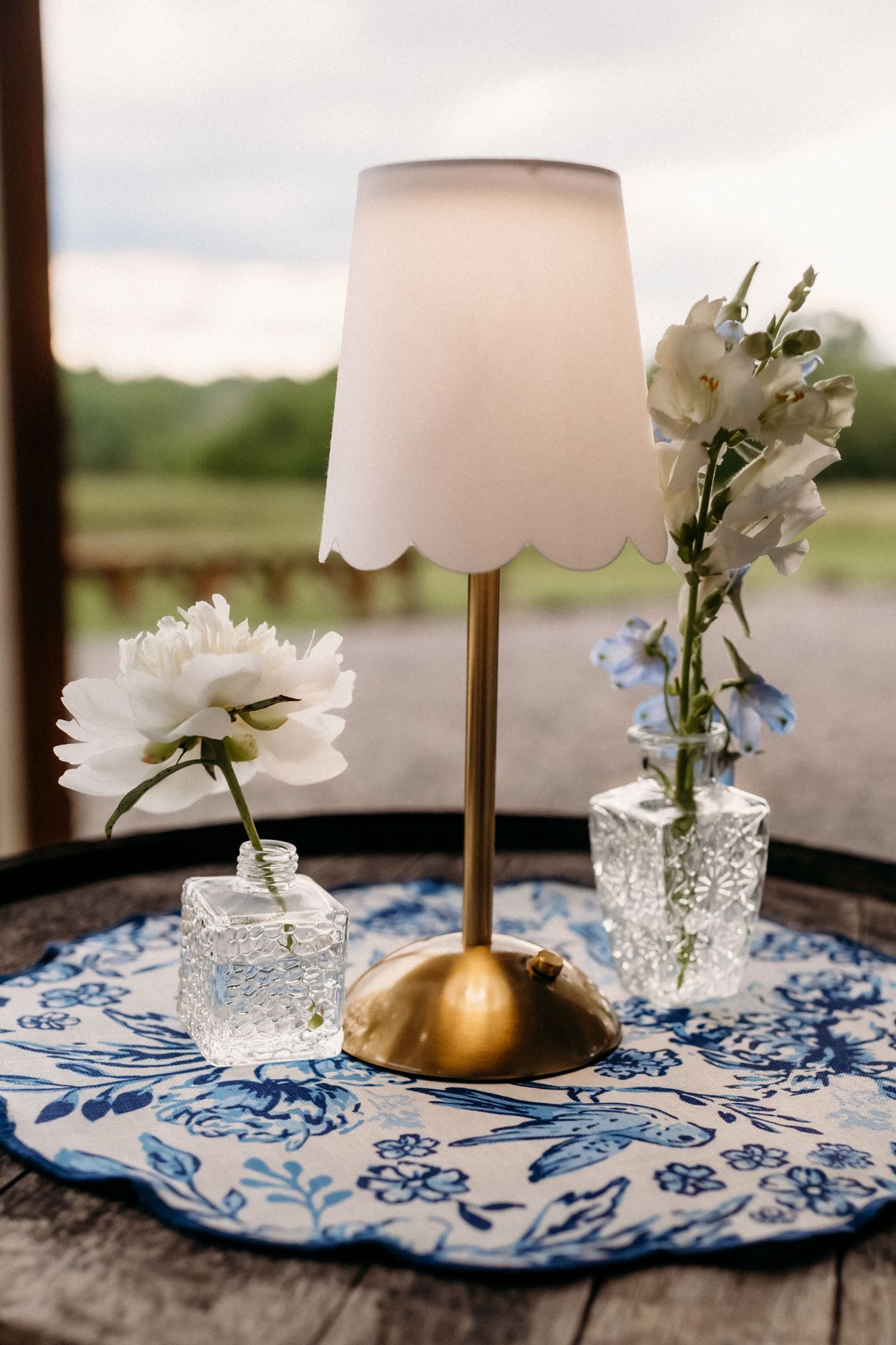 Gold brass table lamp with scalloped shade surrounded by white flowers in crystal vases on blue floral mat.