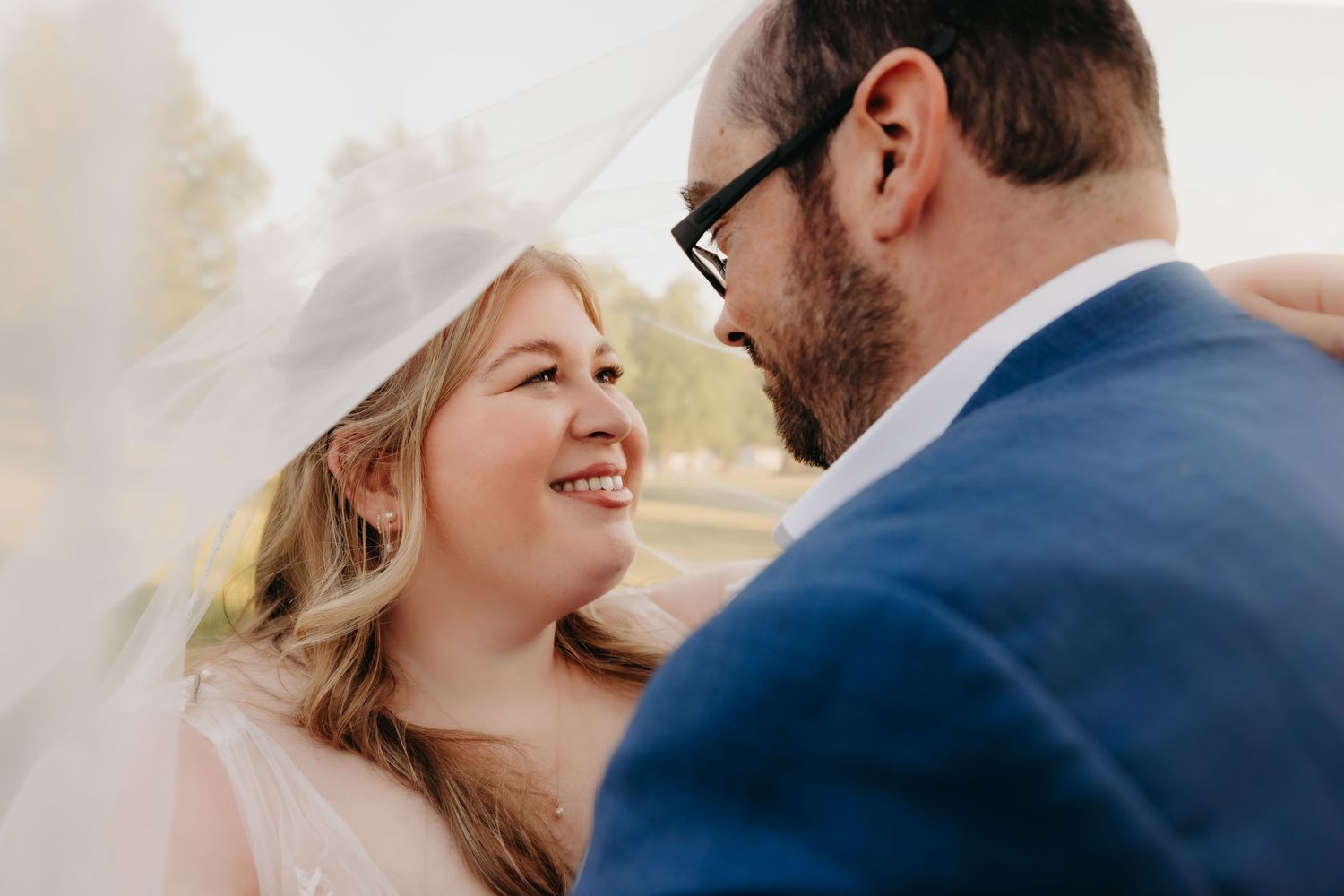 Smiling bride gazes at her groom under a veil in a romantic close-up wedding portrait outdoors.