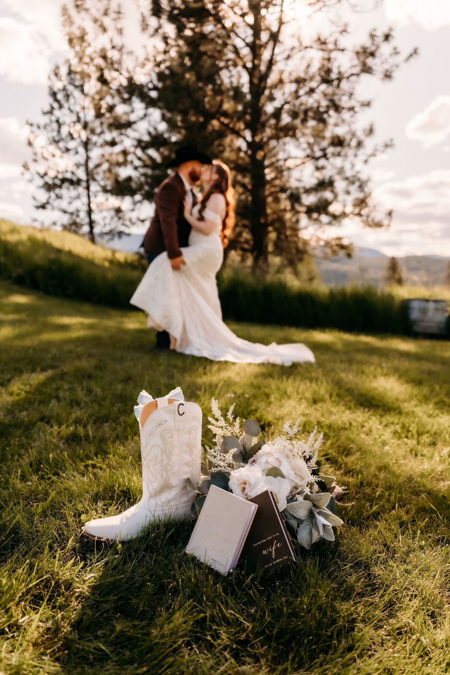 White cowboy boots and bridal bouquet in foreground as couple kisses at golden hour outdoor wedding.