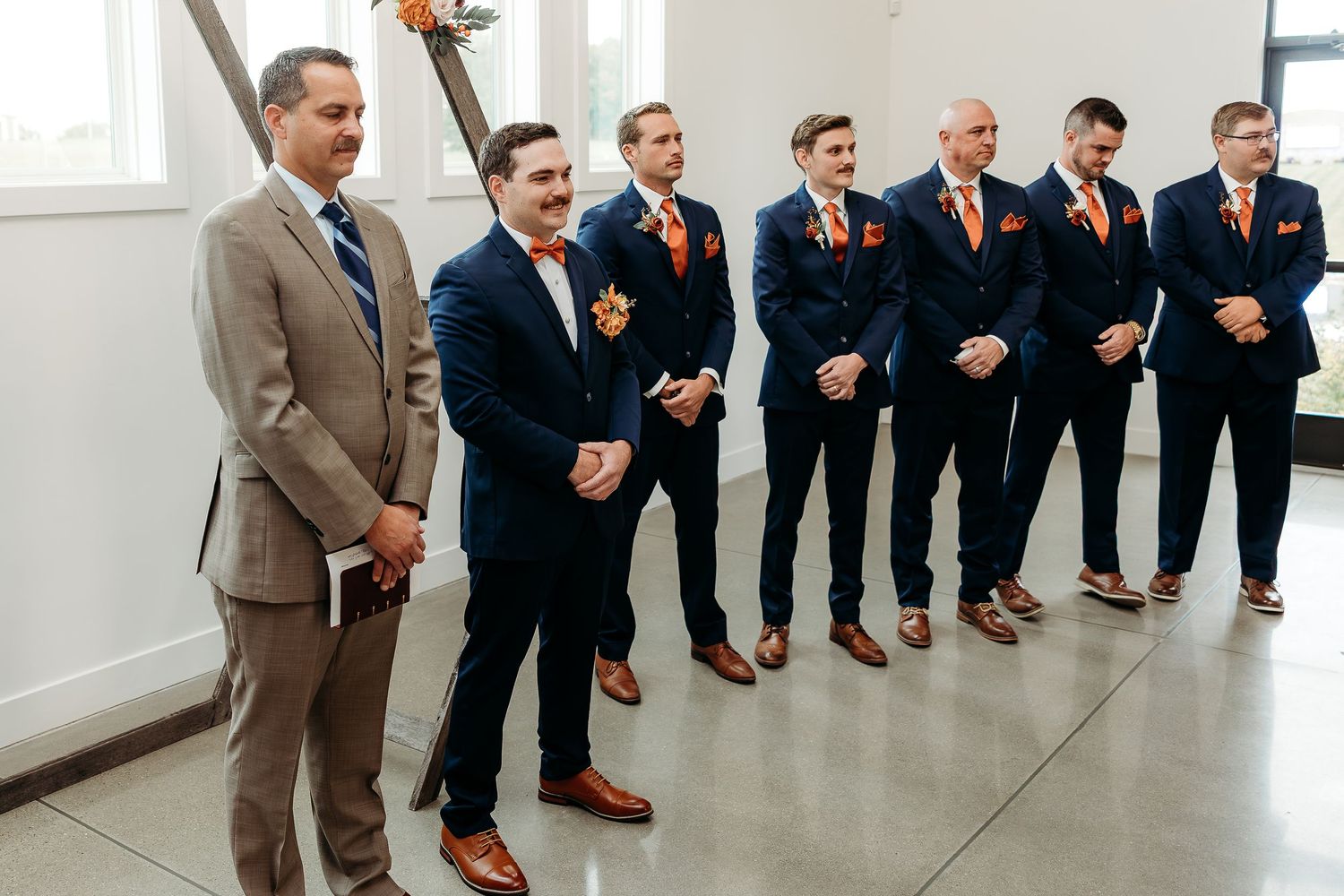 Groom and groomsmen in navy blue suits with orange boutonnieres and ties lined up at a wedding ceremony.