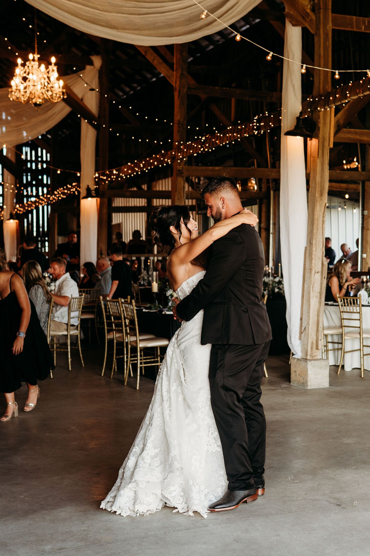 Bride and groom share first dance in a rustic barn venue with string lights and chandeliers.