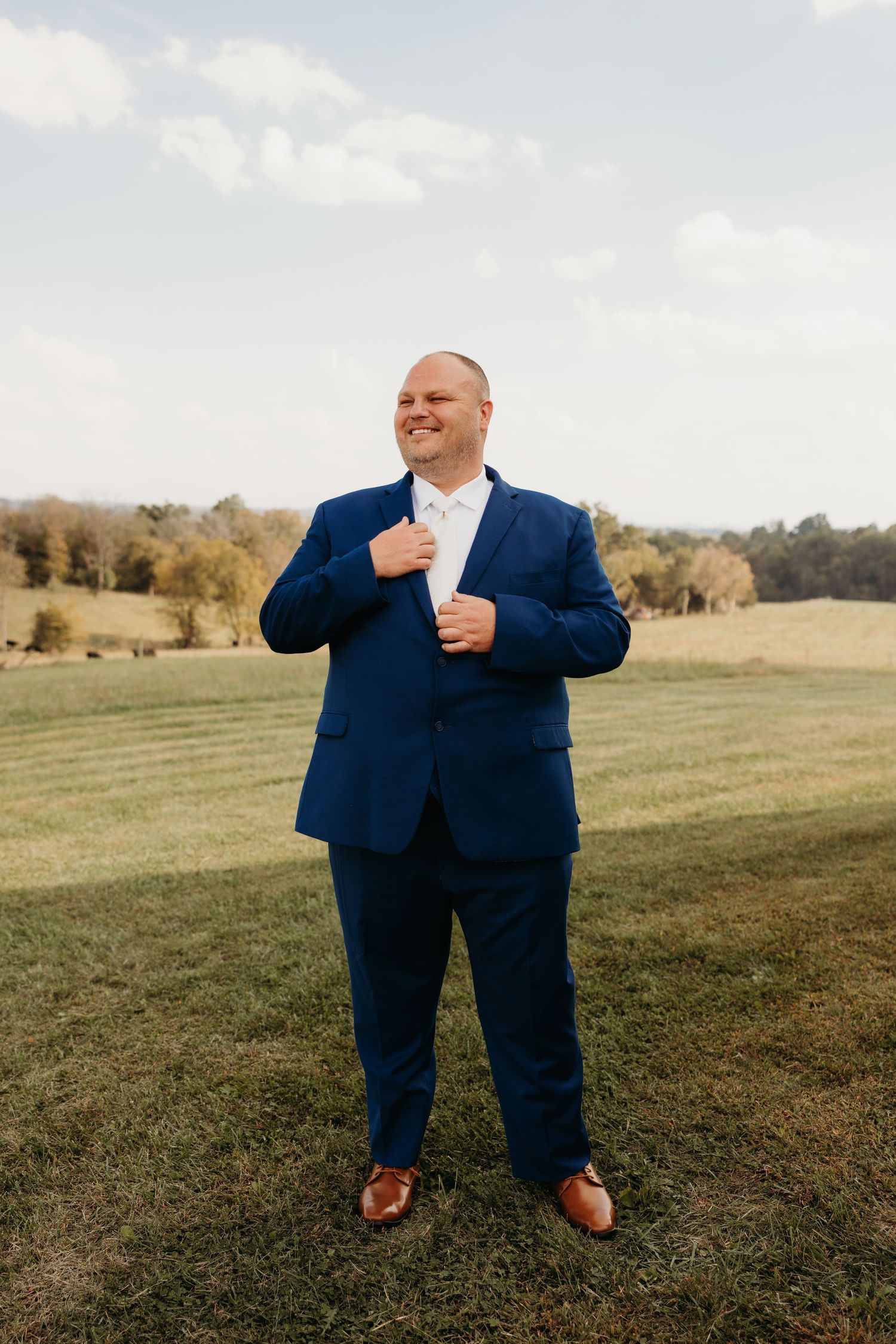 Groom in navy blue suit adjusts jacket while smiling outdoors in a green field on wedding day.