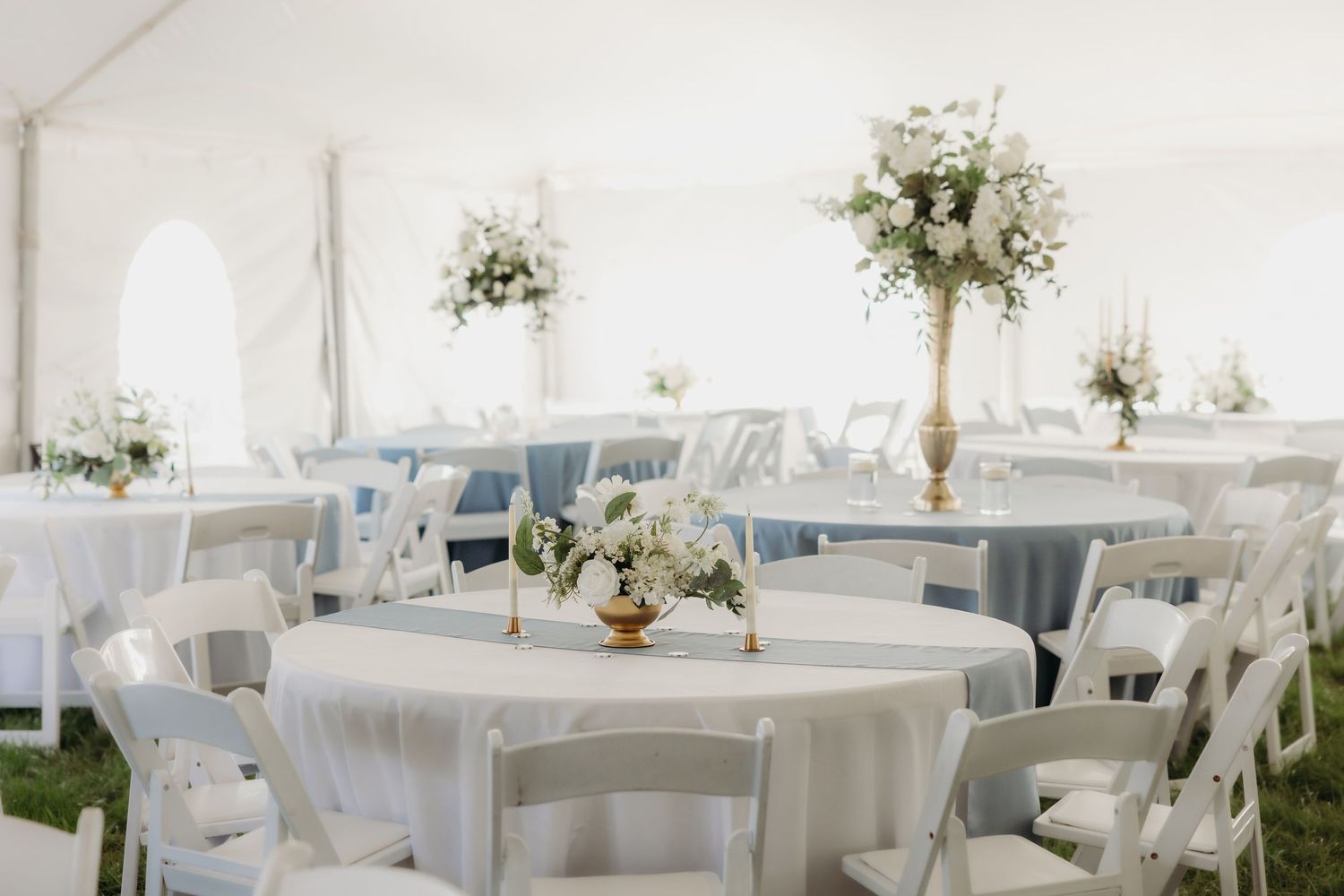 Elegant white wedding reception tent with round tables, white chairs, and tall gold floral centerpieces with white flowers.
