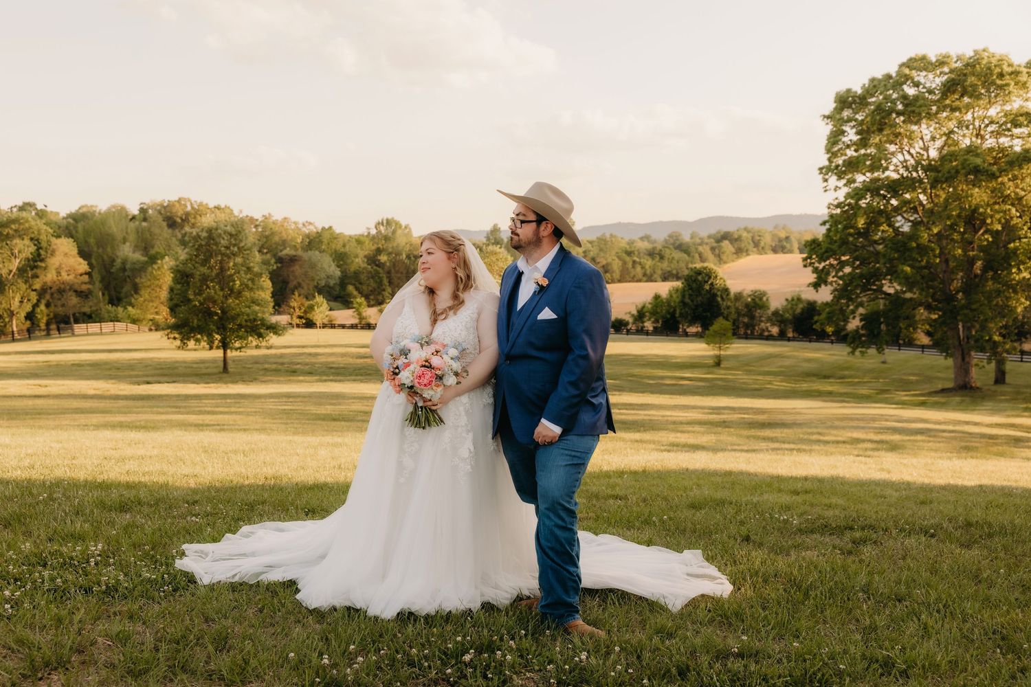 Bride and groom in navy suit and cowboy hat share a romantic moment in a sunlit green field.