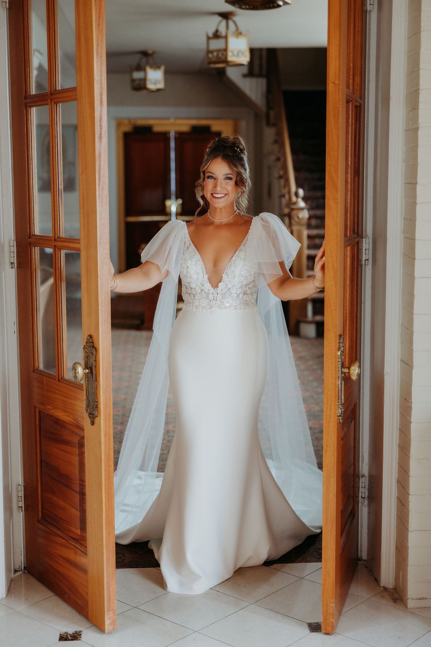 A white wedding dress with lace details hangs in a wooden doorway.