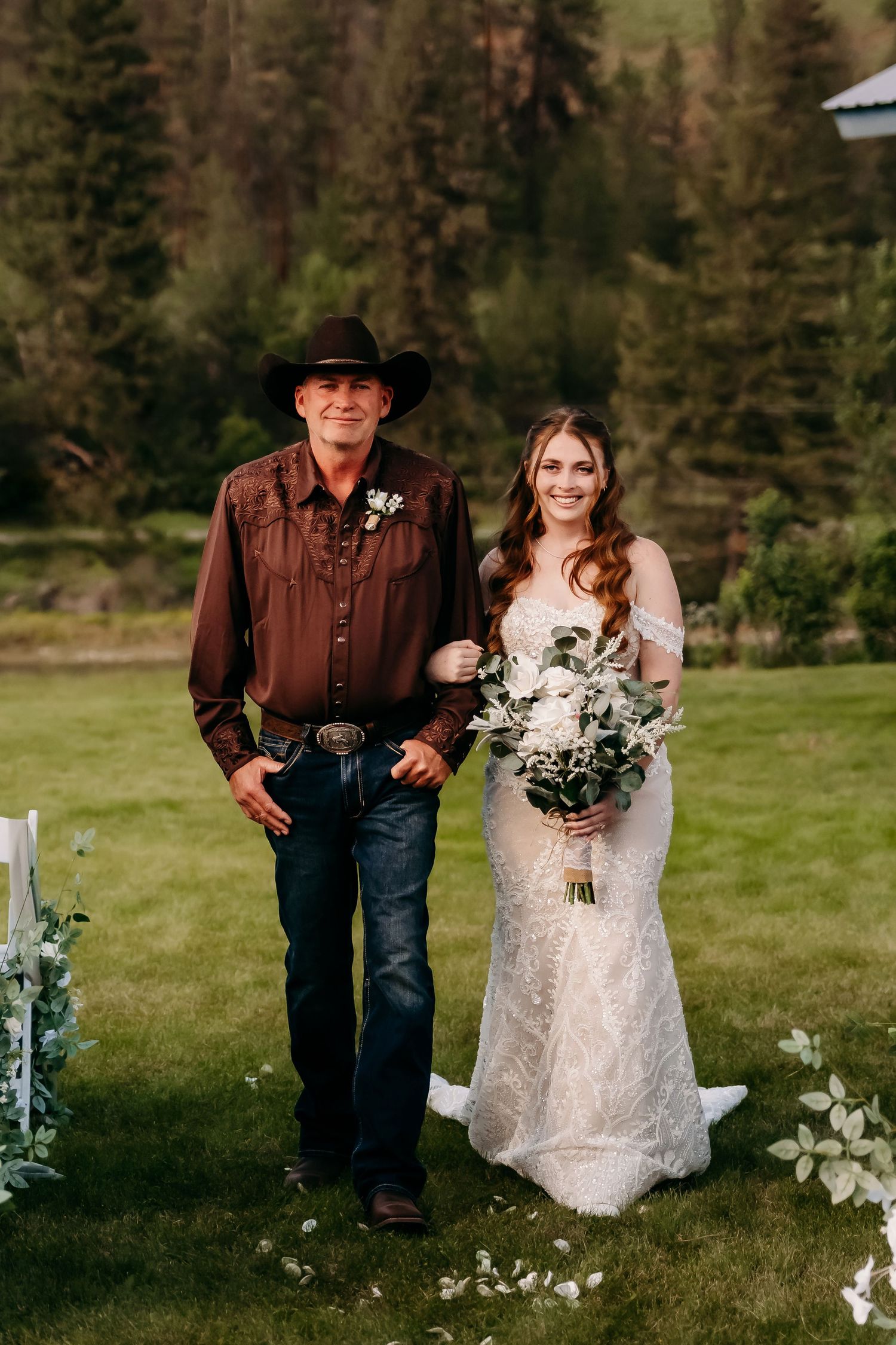 Father walks bride down the aisle outdoors, bride holding white bouquet in lace wedding gown.