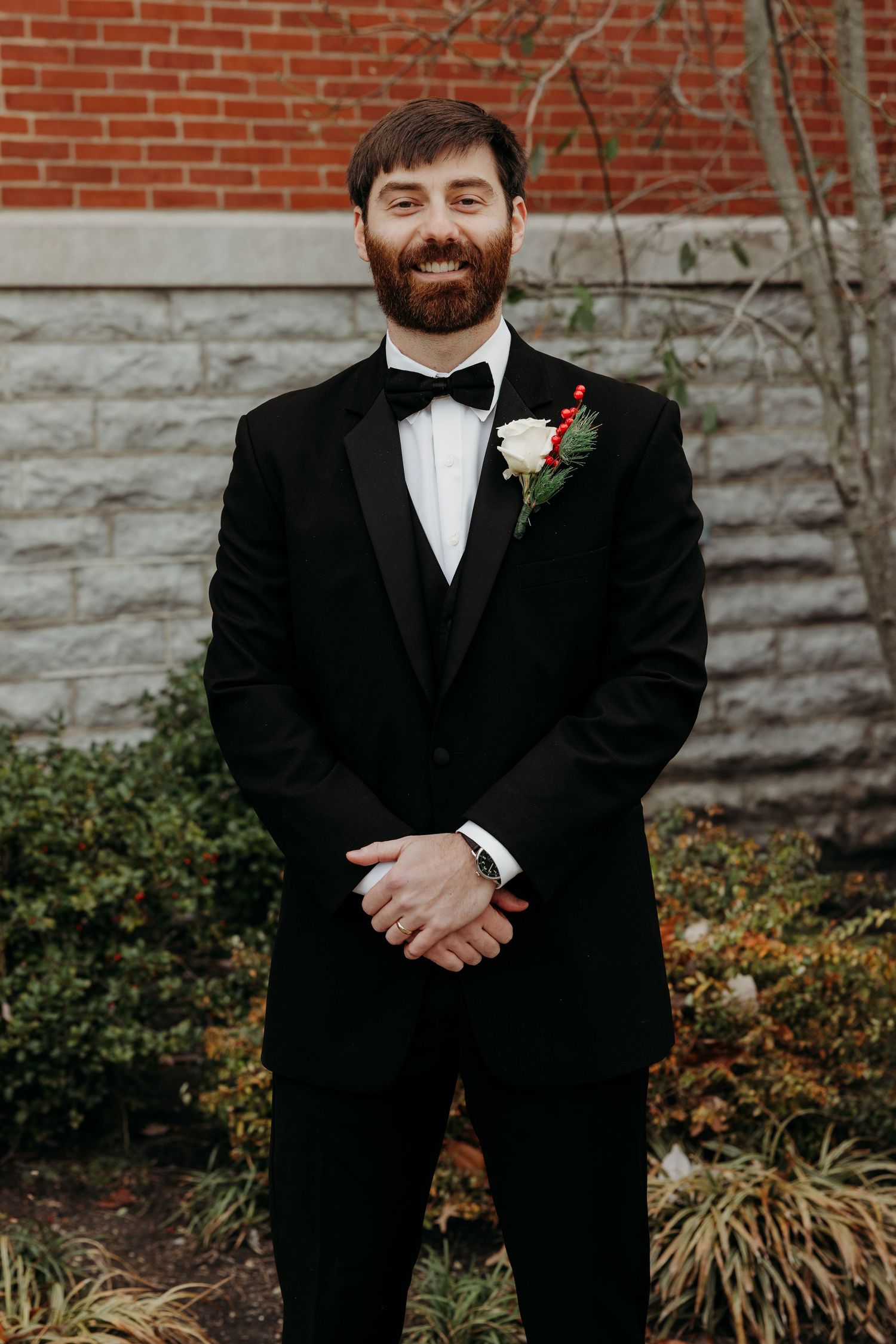 Smiling groom in black tuxedo with bow tie and floral boutonniere poses outdoors against brick wall.