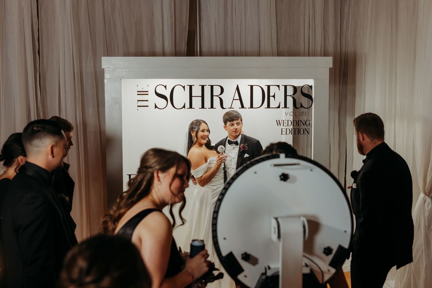 Wedding guests gathered at a magazine-style photo booth backdrop reading The Schraders Wedding Edition.