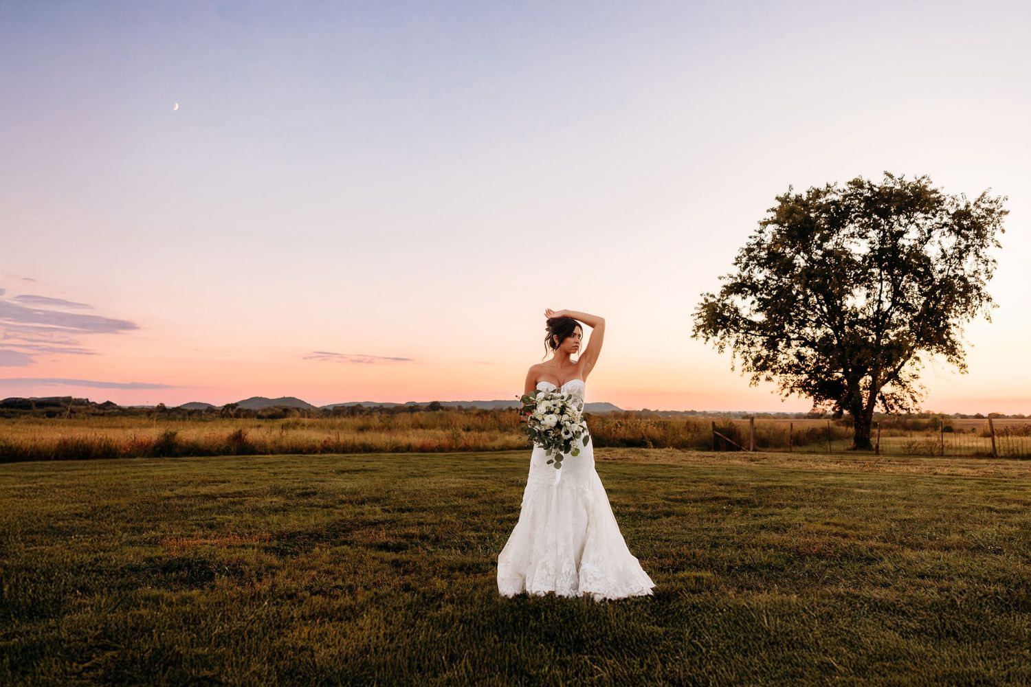Elegant bride holding bouquet gazing at colorful sunset sky beside a tree in an open field.