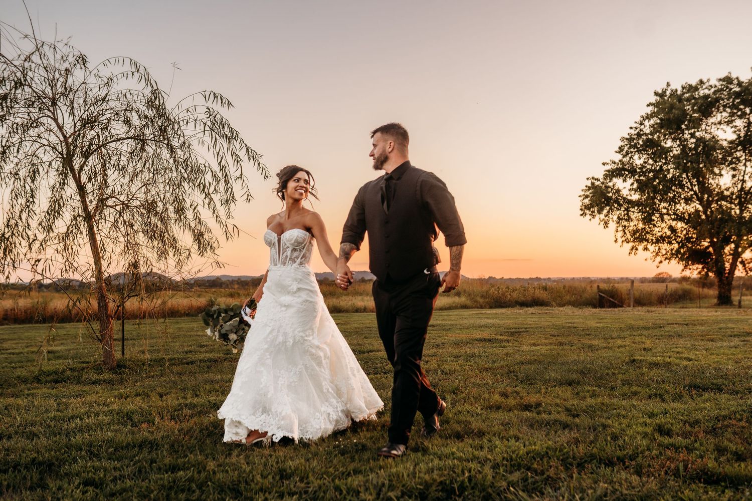Silhouettes of newlyweds dance in a field during golden hour with willow trees in background.