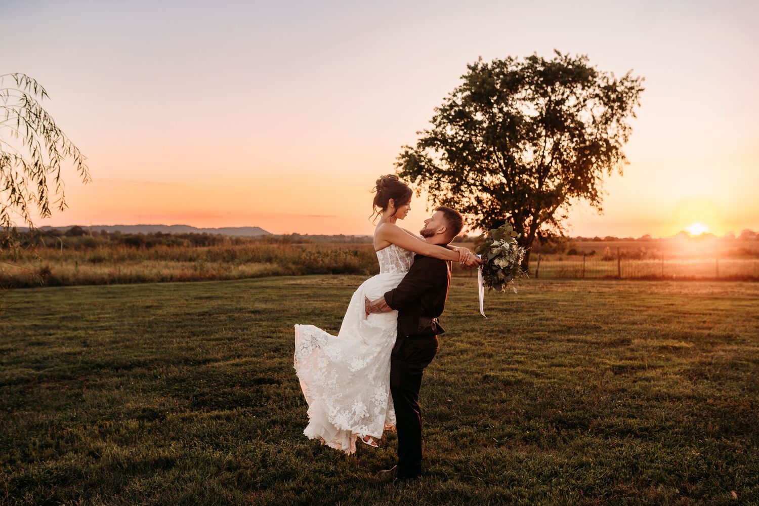 Groom lifting bride in white lace dress at golden sunset near tree in green field.