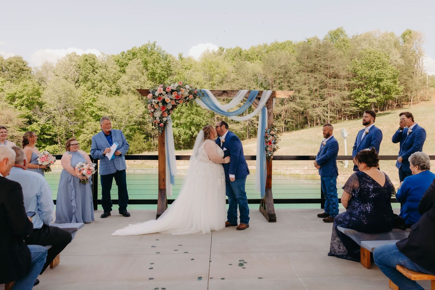 Outdoor wedding ceremony with couple at floral arch, groomsmen in blue suits, guests seated.