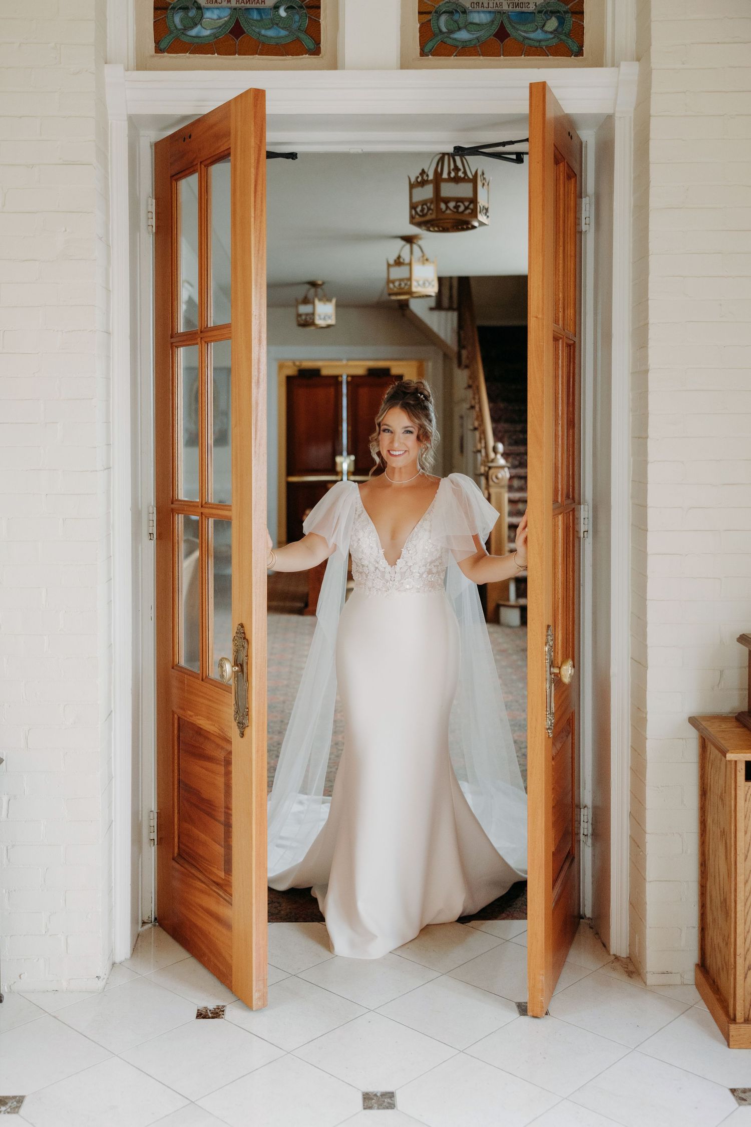 Radiant bride in lace flutter-sleeve wedding gown stands in open wooden double doors of elegant venue.