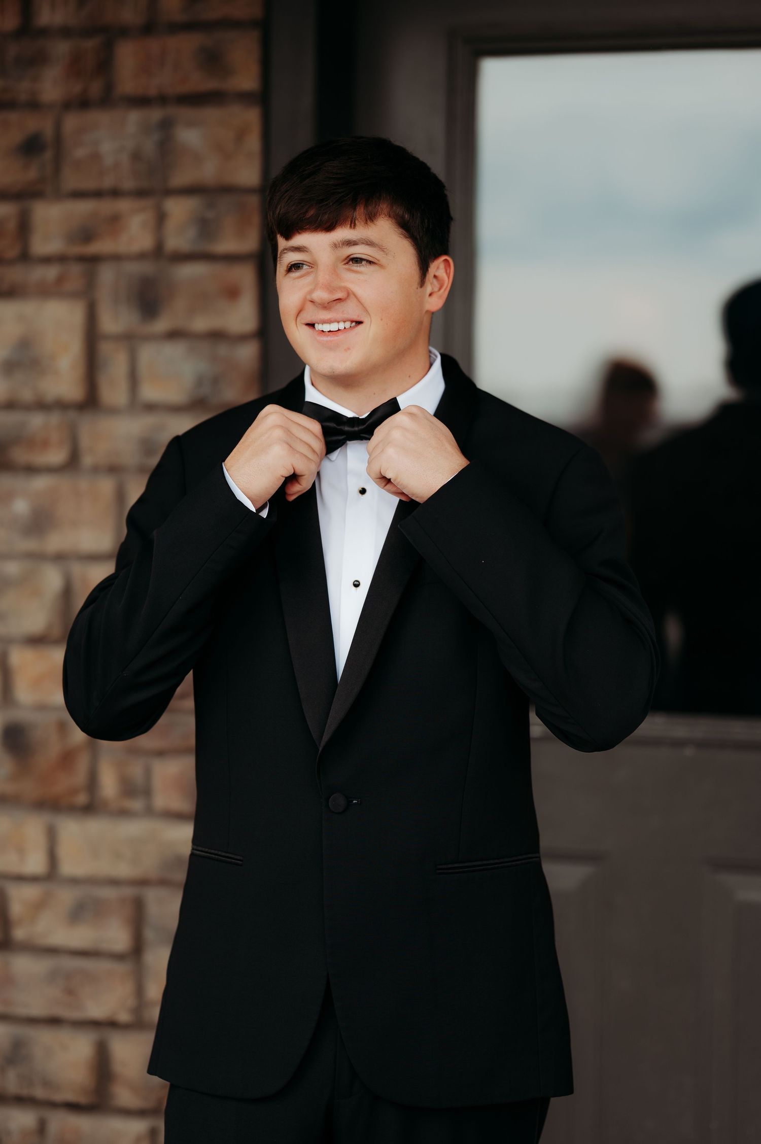 Happy groom in black tuxedo adjusting bow tie while standing in front of a brick wall.