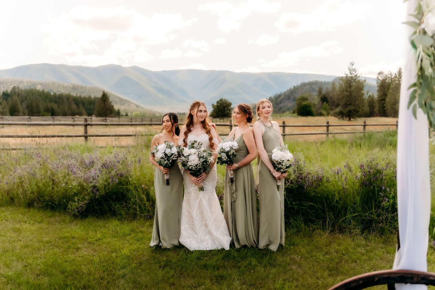 Bride with three bridesmaids in sage green gowns holding bouquets with mountain valley backdrop outdoors.