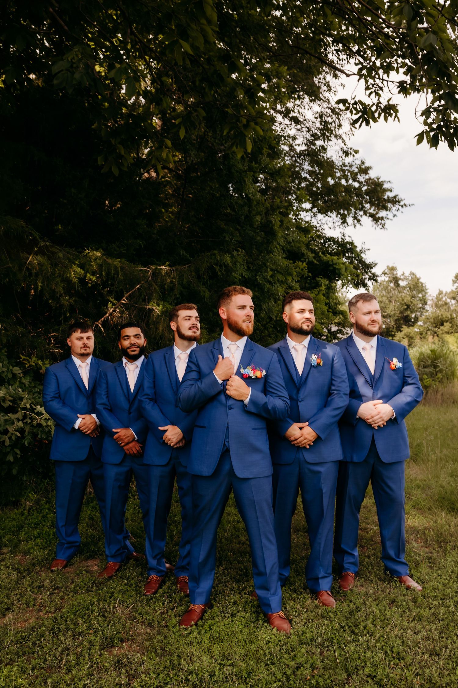 Groom and six groomsmen in matching royal blue suits posing outdoors under green trees.