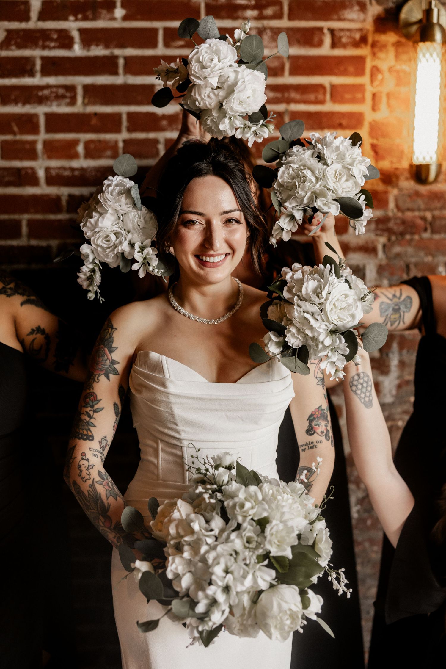 A wedding bouquet of white roses and greenery held against a rustic brick wall with warm lighting.