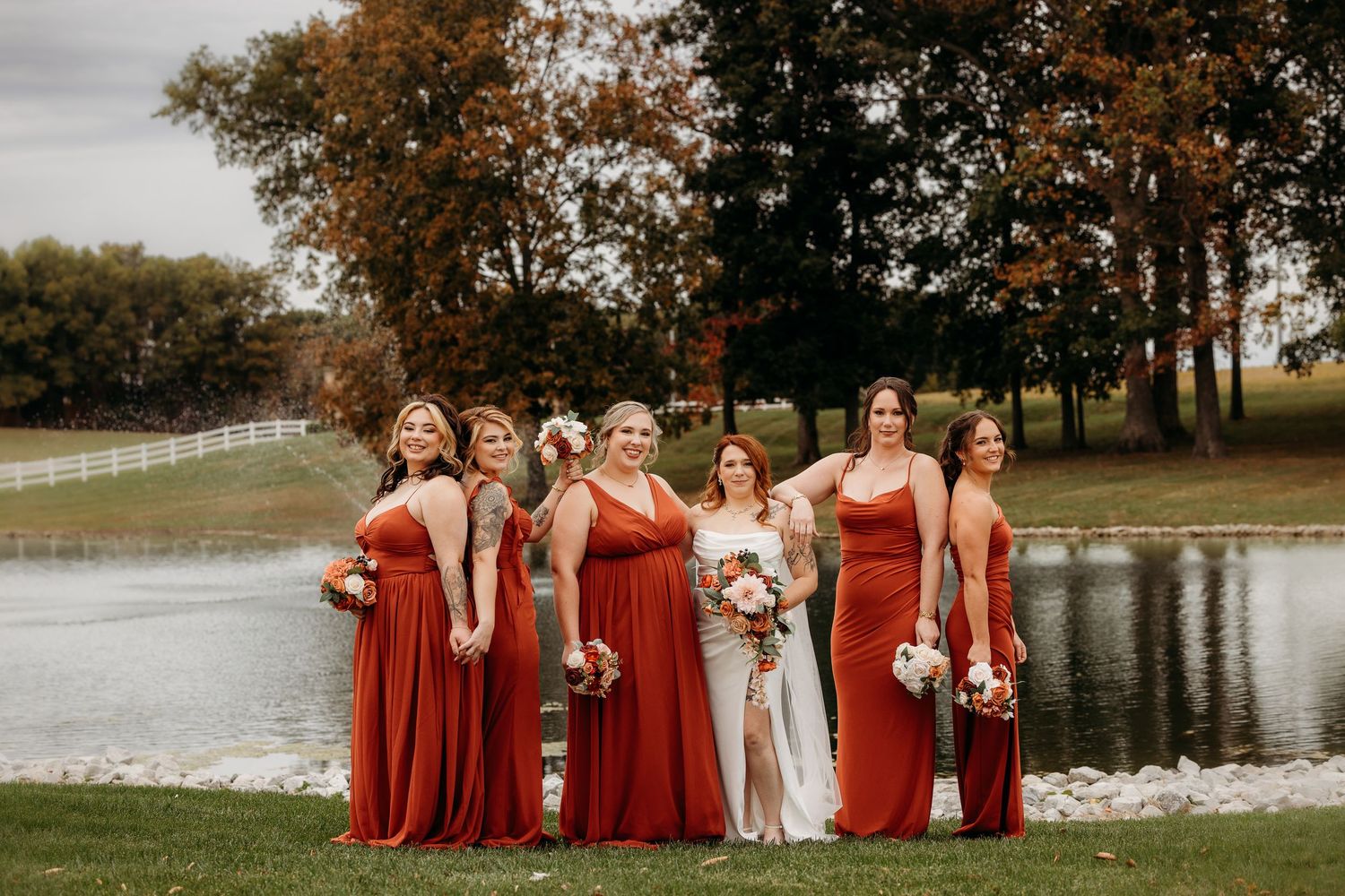 Bridal party in rust-colored dresses poses by a lake during autumn.