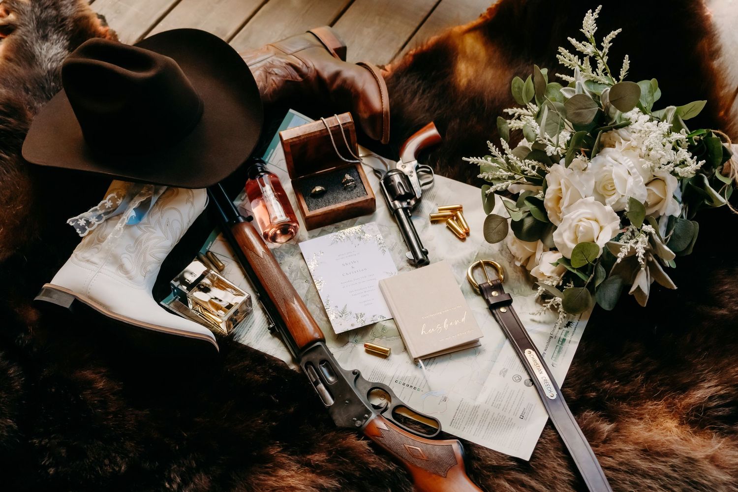 Rustic wedding flat lay with rifle, revolver, cowboy hat, white floral bouquet, and stationery on fur.