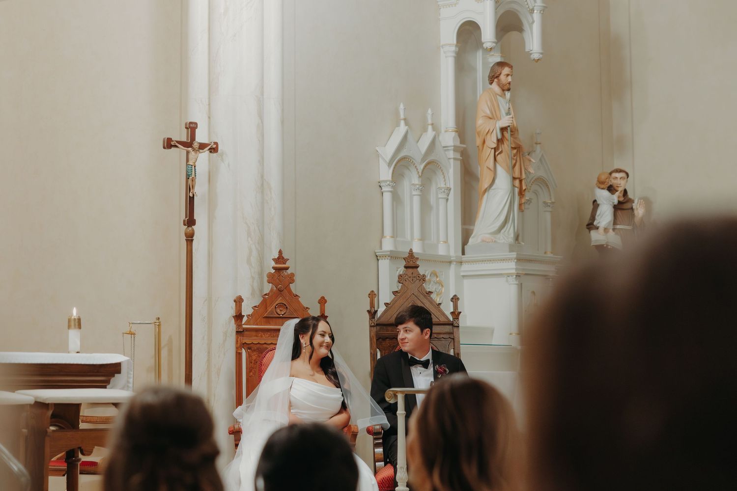 Bride and groom seated at the altar during a Catholic church wedding ceremony with ornate white decor.