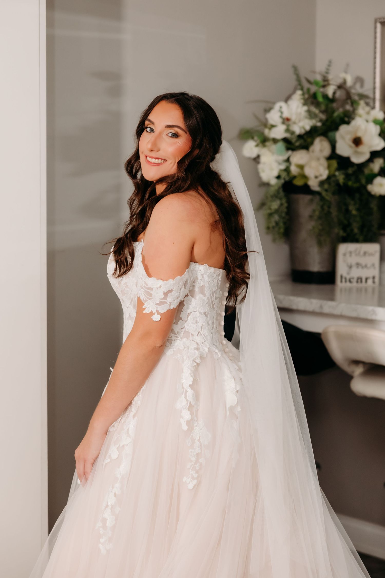 Bride in off-shoulder lace wedding gown with veil smiling indoors.