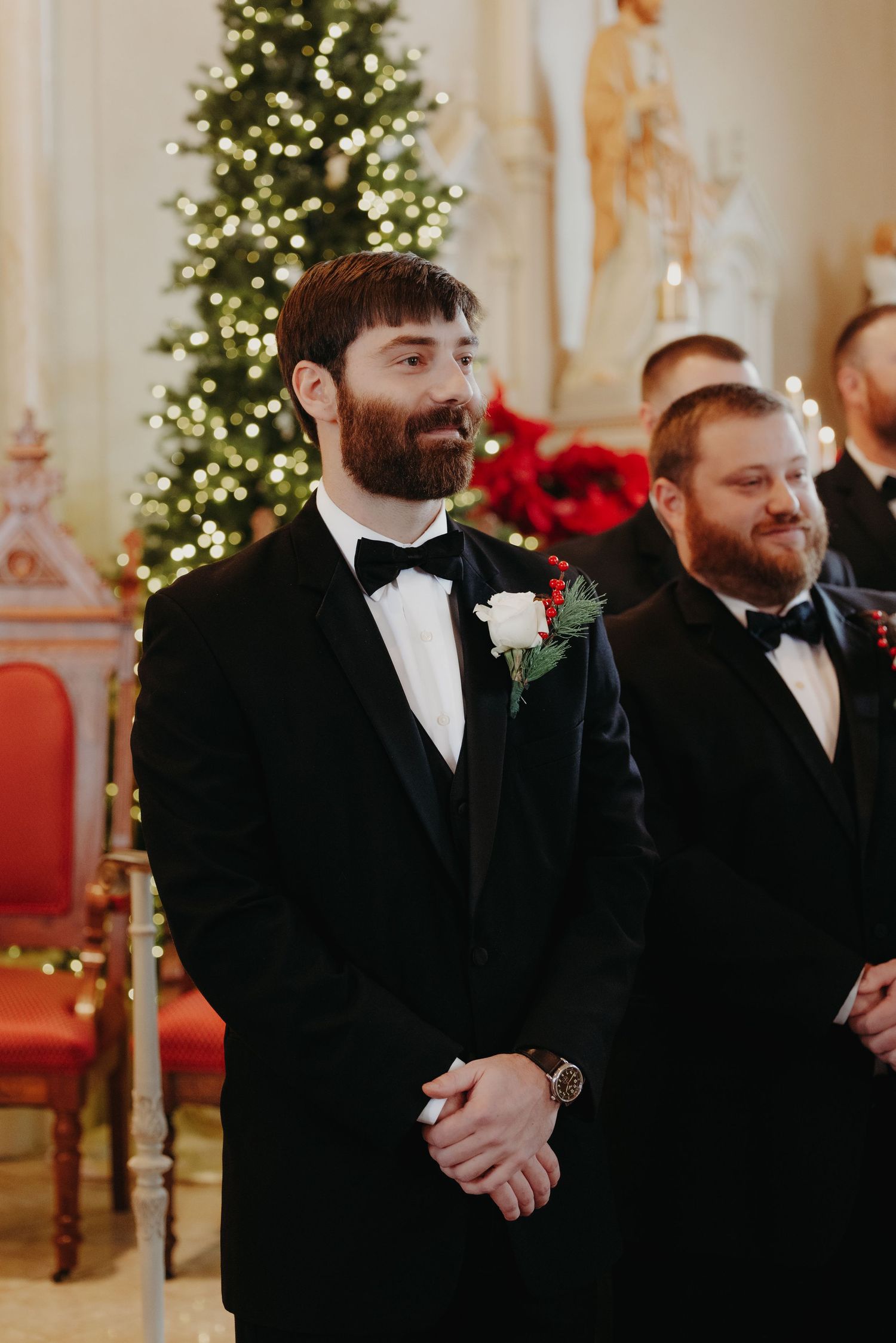 Bearded groom in black tuxedo with white rose boutonniere smiles at altar beside Christmas tree in church.