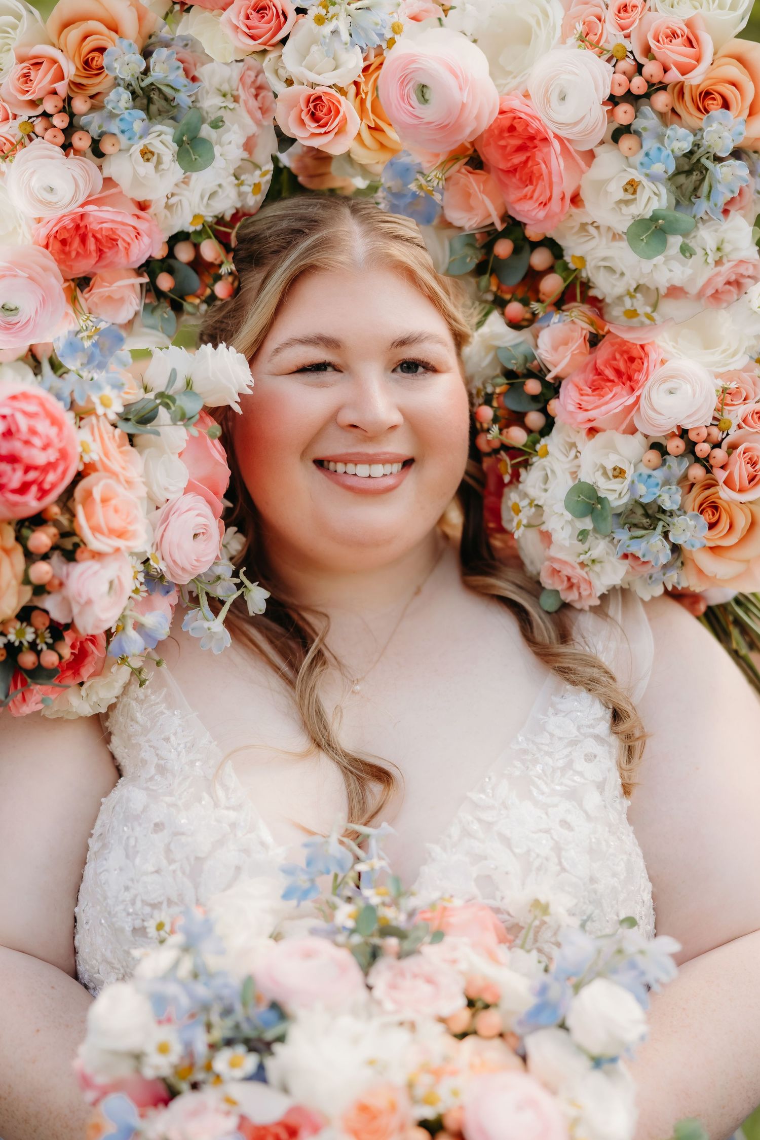 Smiling bride in lace wedding dress lying among colorful pink, peach, and blue flowers holding a bouquet.