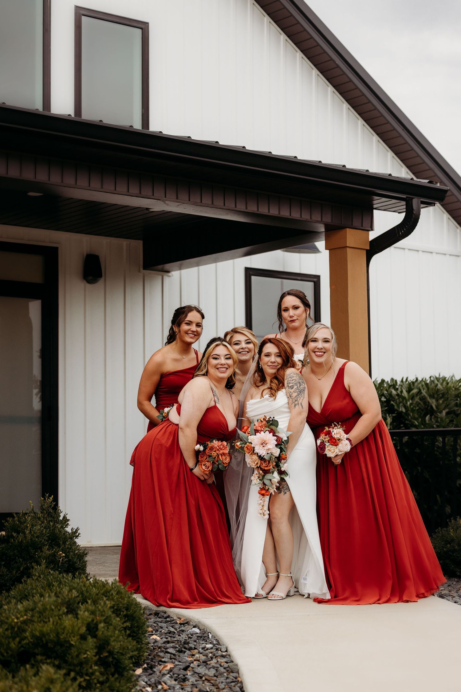 Bride in white gown poses with four bridesmaids in red dresses holding bouquets outside a modern white barn venue.