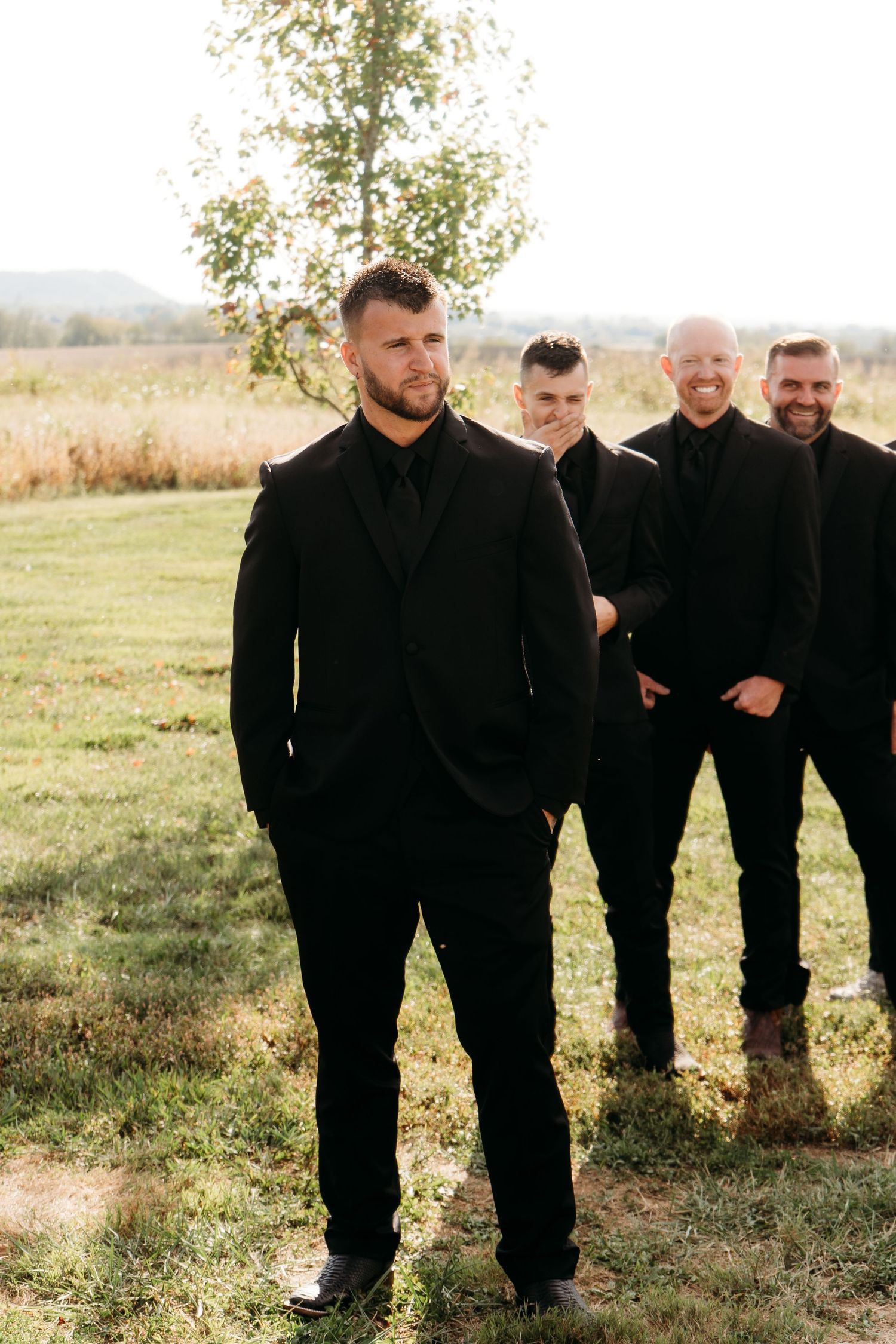 Four groomsmen in matching all-black suits stand outdoors in a field, with the groom positioned prominently in the foreground.
