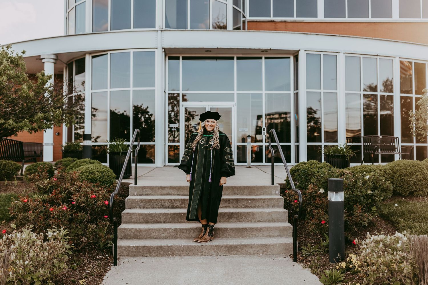 College graduate posing in front of architectural view of a contemporary building entrance with stone steps and glass facade.