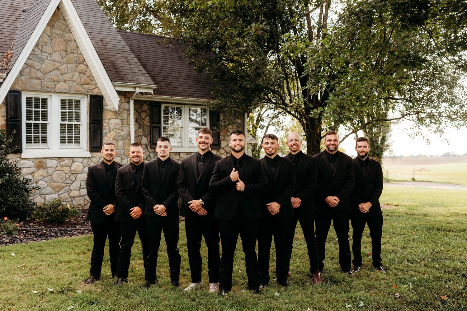 Nine groomsmen in matching all-black suits pose together on a lawn in front of a stone cottage.