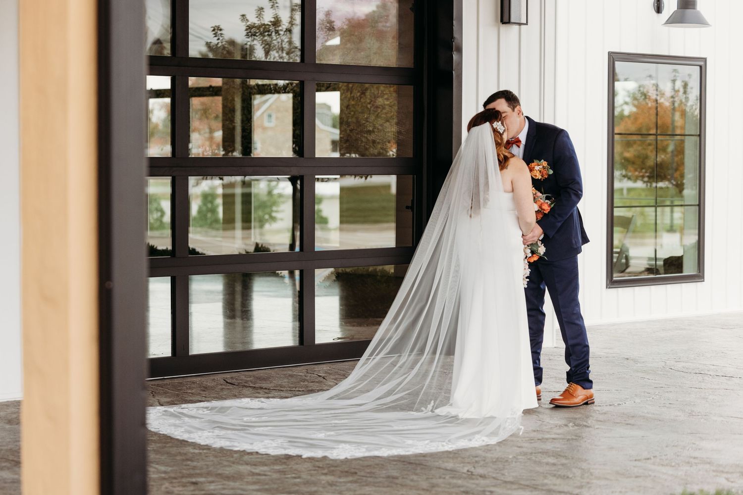 Bride and groom share a romantic kiss outside a modern venue, her cathedral veil trailing elegantly on the ground.