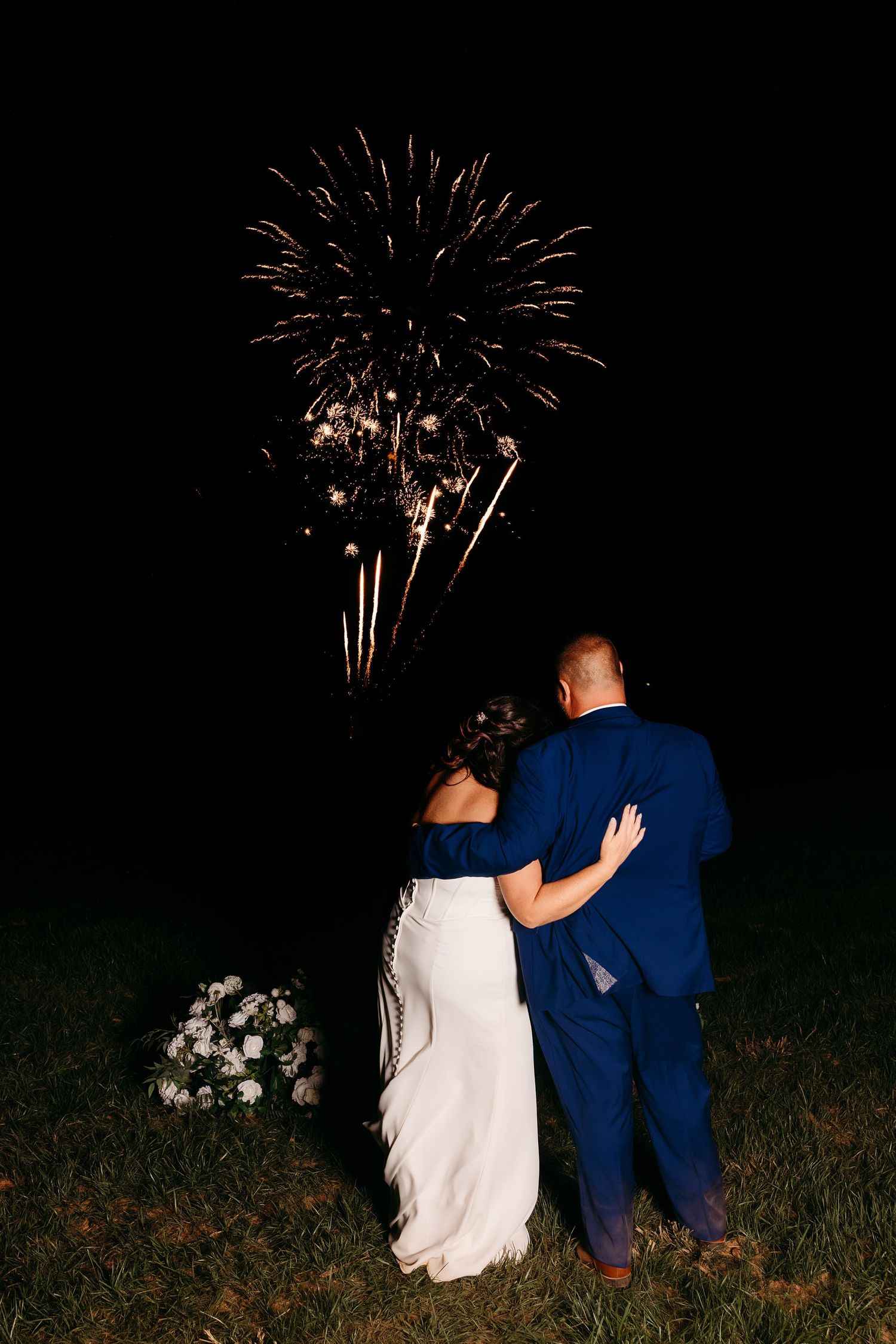 Newlywed couple embraces while watching colorful fireworks light up the night sky at their wedding celebration.