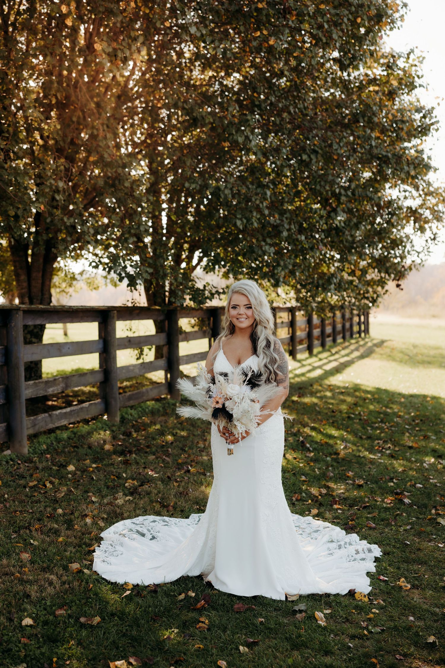 Bride in fitted white lace gown holds boho bouquet beside wooden fence at fall farm venue.