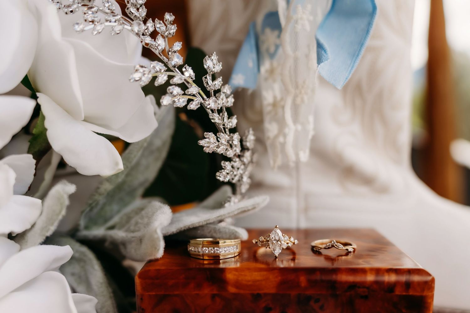 Wedding rings and diamond hair piece displayed on wooden box beside white floral bouquet and bridal gown.