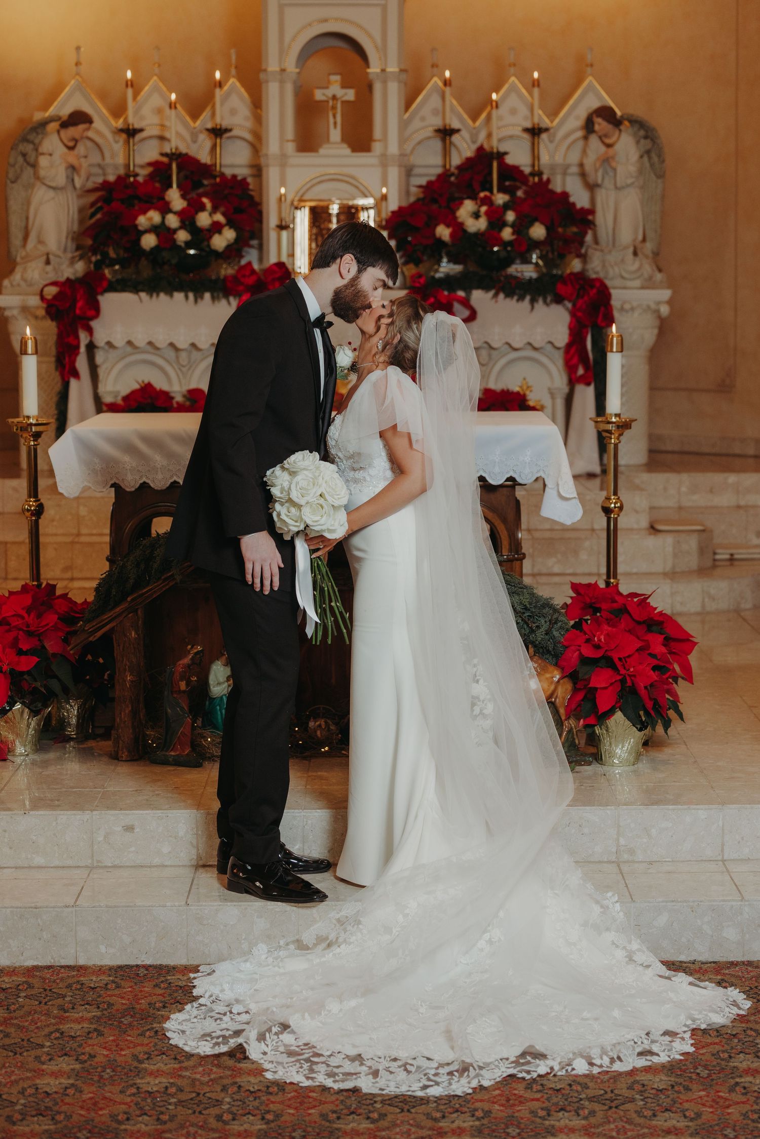 Bride and groom share first kiss at altar adorned with red poinsettias and candles during Christmas wedding ceremony.