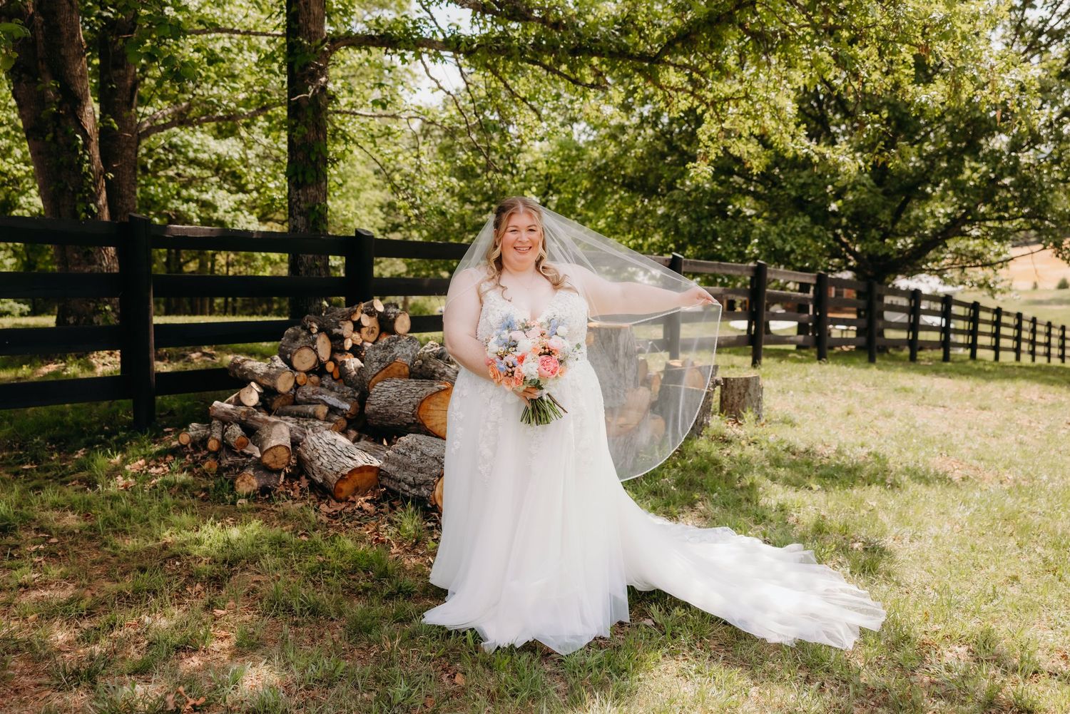 Bride in white gown and veil holding bouquet outdoors near log pile and wooden fence.