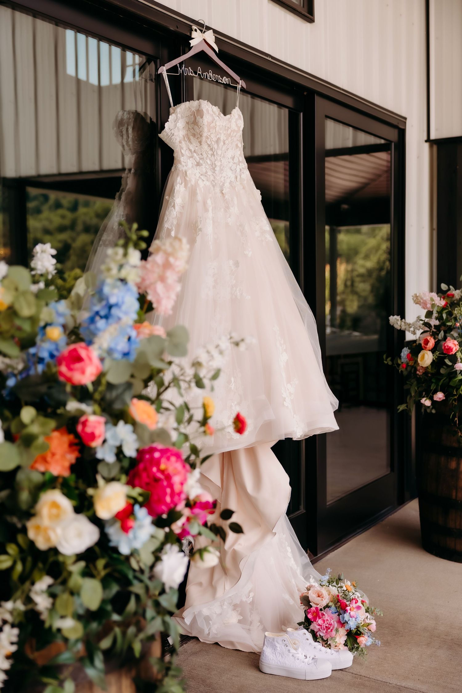 Elegant lace wedding dress hanging on personalized hanger with colorful floral arrangements and white Converse shoes below.