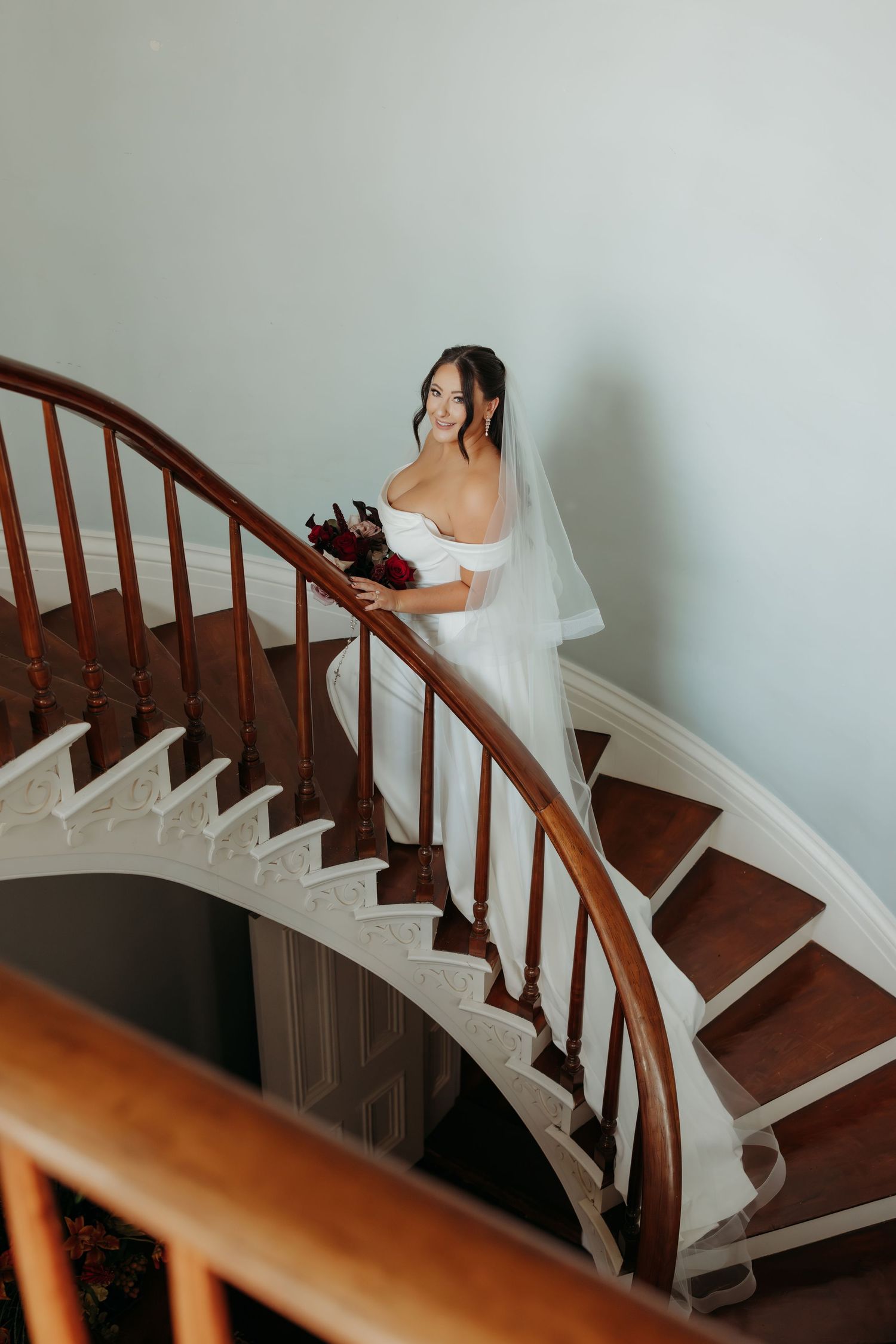 A bride in a white wedding dress descends an elegant curved wooden staircase while holding a bouquet at Simmstown Manor wedding venue.
