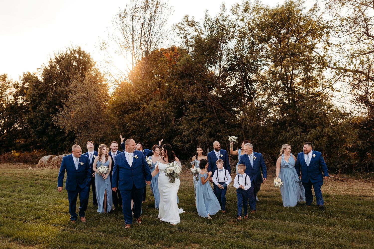 Wedding party in blue attire walking and laughing together in golden sunset light.
