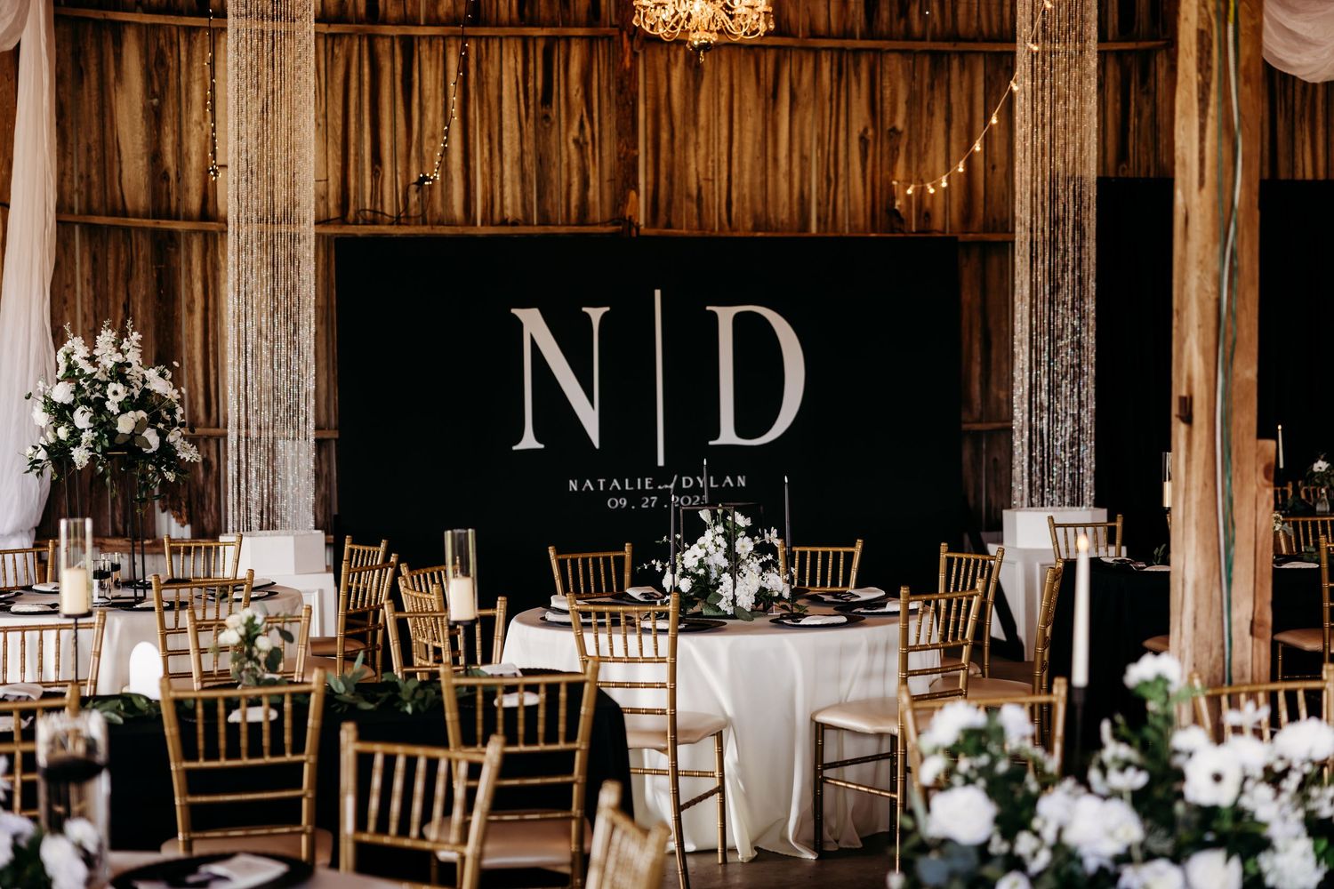 Elegant barn wedding reception with gold chiavari chairs, white floral centerpieces, and monogram ND backdrop.