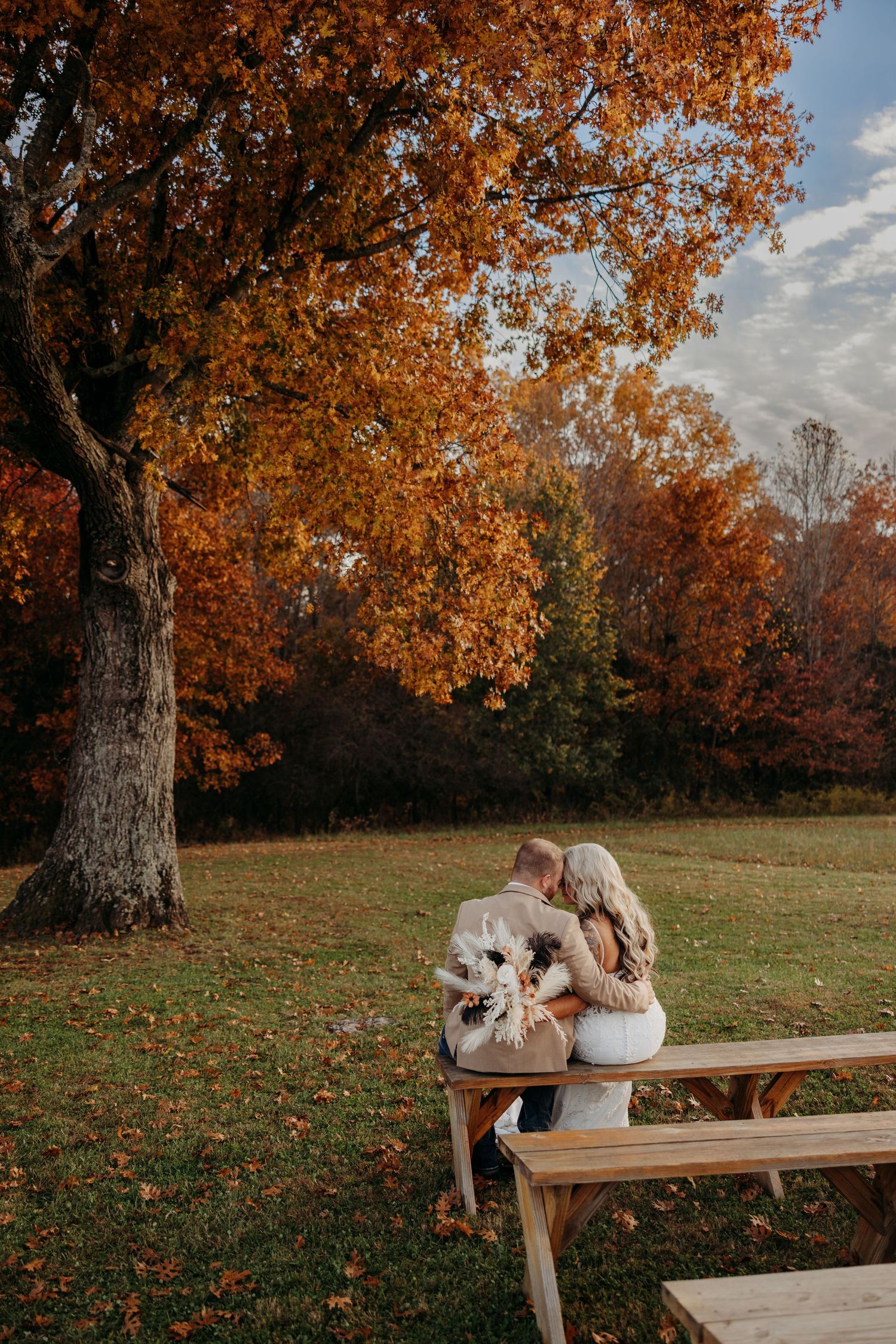 Couple shares romantic moment on bench beneath vibrant orange autumn trees holding wedding bouquet.