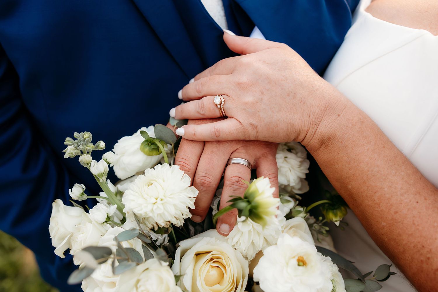 Bride and groom hands with wedding rings resting on white rose bouquet at wedding ceremony.