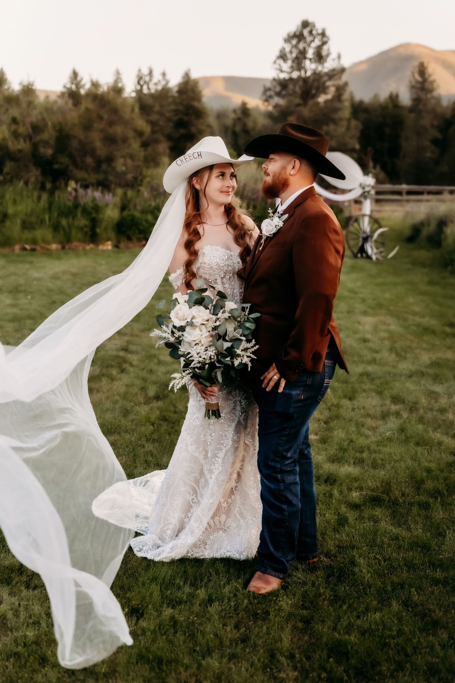 Western wedding couple in cowboy hats share romantic moment on green lawn with mountains and flowing bridal veil.